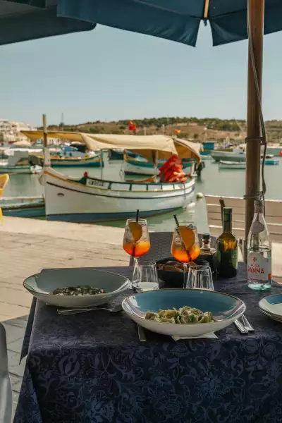Malta | Two Aperol spritzes sit on an outdoor table laden with food, with a docked boat in the background in Malta