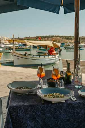 Malta | Two Aperol spritzes sit on an outdoor table laden with food, with a docked boat in the background in Malta