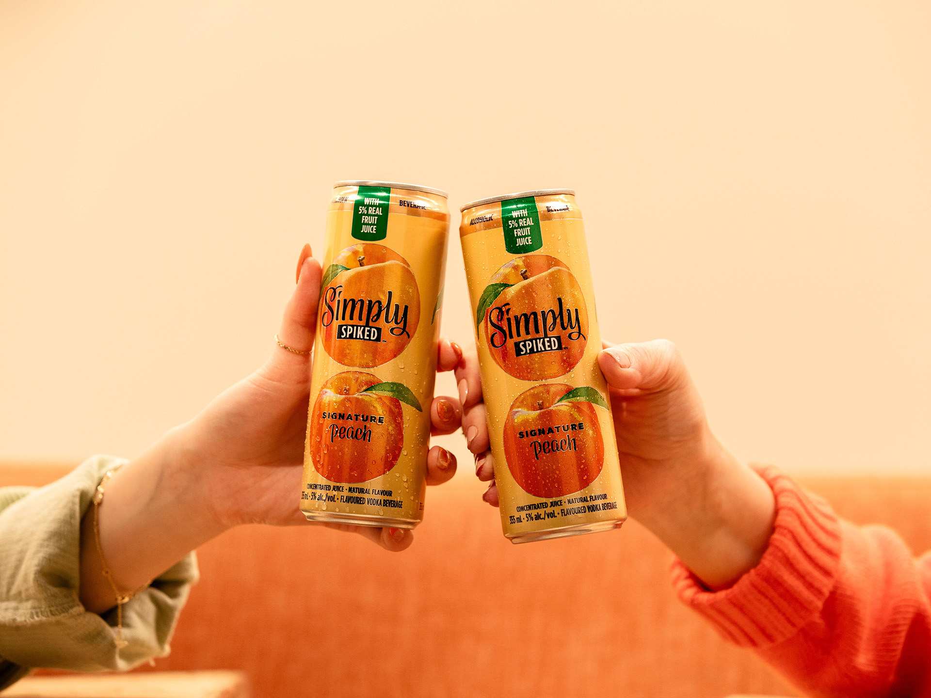 Two people cheers-ing with Simply Spiked peach cans Two people cheers-ing with Simply Spiked peach cans
