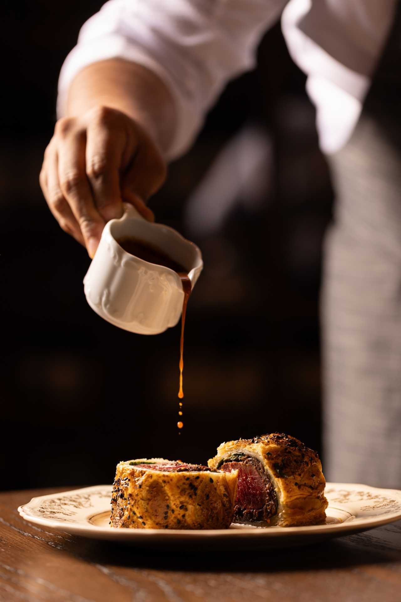 Punch Toronto | A hand pours a rogan josh jus over the Beef Wellington at Punch